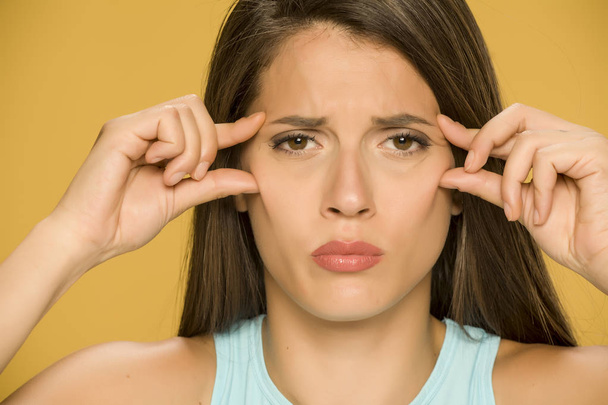 Young worried woman pinching her eye wrinkles on yellow background - Photo, Image
