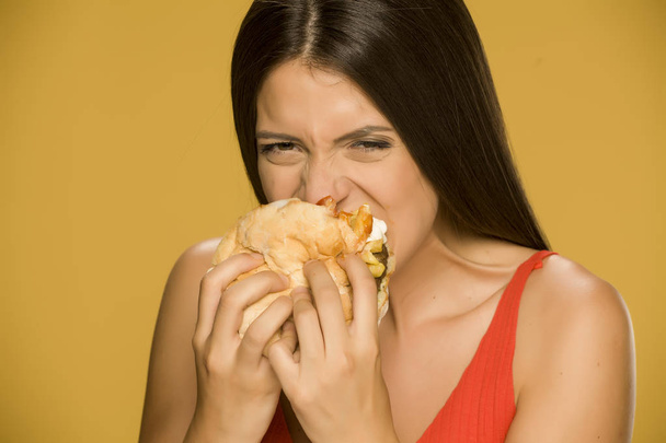 Jonge hebzuchtige vrouw het eten van een hamburger op gele achtergrond - Foto, afbeelding