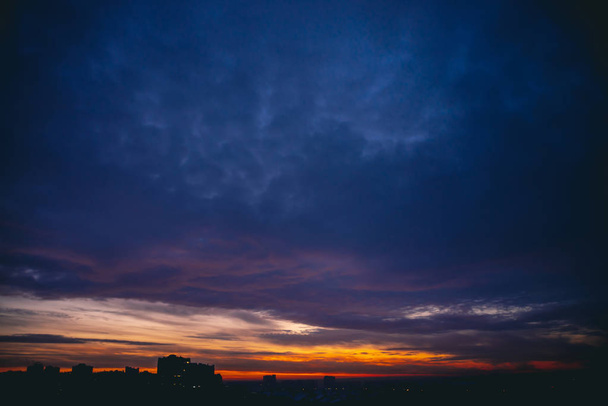Cityscape com vívida alvorada quente. Espantoso céu azul violeta nublado acima de silhuetas escuras de edifícios da cidade. Luz solar laranja. Fundo atmosférico do nascer do sol em tempo nublado. Espaço de cópia
. - Foto, Imagem