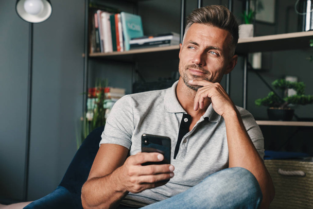 Photo of happy caucasian man holding smartphone while sitting on armchair at home - Foto, afbeelding