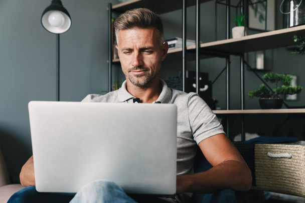 Photo of pleased adult man using laptop while sitting on armchair in apartment - Photo, image