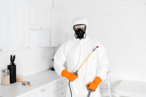 exterminator in protective mask and uniform holding toxic equipment near kitchen cabinet  - Zdjęcie, obraz