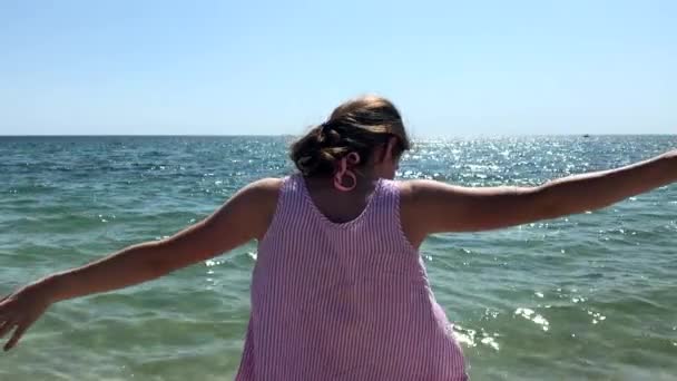 Vista lateral de cerca de una joven mujer caucásica de pie en una playa con sus brazos en el aire, mirando al mar
 - Metraje, vídeo