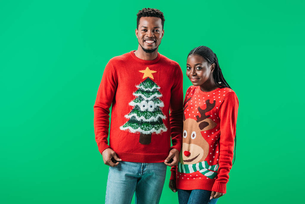 African American man in de buurt van vrouw in rode kerst truien glimlachen en kijken naar camera geïsoleerd op groen - Foto, afbeelding