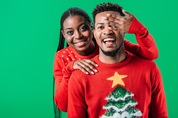 African American woman in Christmas sweater holding hand near eyes of man isolated on green - Foto, immagini