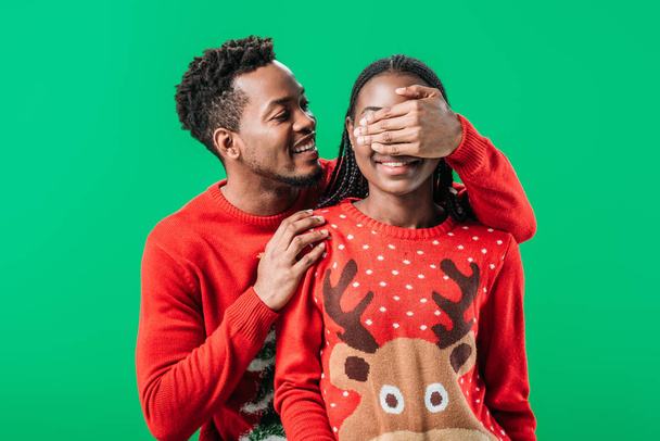 赤いセーターを着たアフリカ系アメリカ人男性が、緑色で孤立した女性に手で目を覆う - 写真・画像