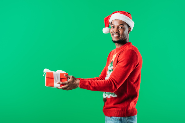 side view of African American man in Christmas sweater and Santa hat holding red gift box looking at camera isolated on green - Fotografie, Obrázek