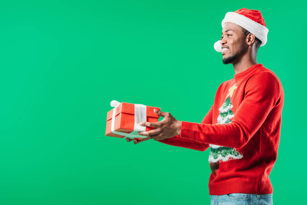 Seitenansicht eines afrikanisch-amerikanischen Mannes in Weihnachtspullover und Weihnachtsmütze mit rotem Geschenkkarton, der isoliert auf grünem Grund wegschaut - Foto, Bild