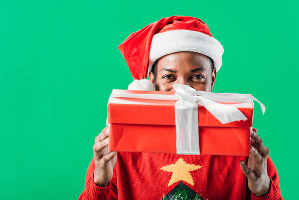 Hombre afroamericano en Santa Sombrero sosteniendo caja de regalo roja con cinta blanca y mirando a la cámara aislada en verde
 - Foto, imagen