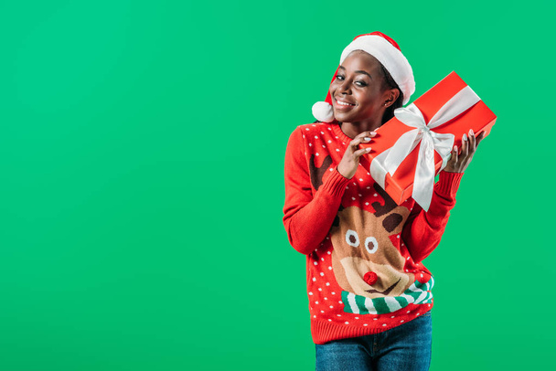 African American woman in Christmas sweater and Santa hat holding gift box and looking at camera isolated on green - Фото, изображение