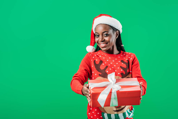 African American woman in Christmas sweater and Santa hat holding gift box with white ribbon and looking at camera isolated on green - Photo, Image