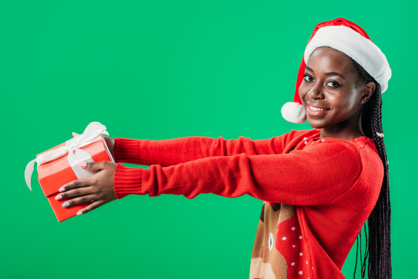 side view of African American woman in Christmas sweater and Santa hat raising hands with gift box and looking at camera isolated on green - 写真・画像