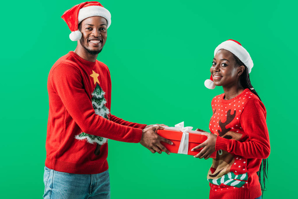 πλευρά του αφρικανικού άνδρα με κόκκινο χριστουγεννιάτικο πουλόβερ τραβώντας το κουτί δώρου με γυναίκα απομονωμένη στο πράσινο - Φωτογραφία, εικόνα