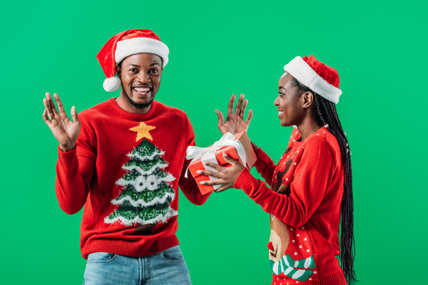 African American woman in red Christmas sweater and Santa hat giving gift box to surprised man isolated on green - Foto, immagini