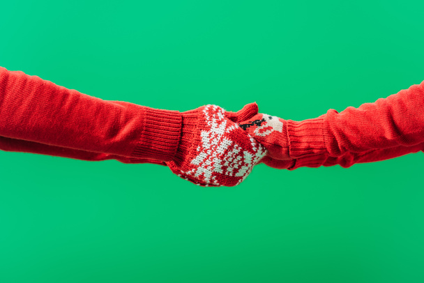 bijgesneden beeld van twee mensen in rode truien houden handen in de winter handschoenen geïsoleerd op groen - Foto, afbeelding