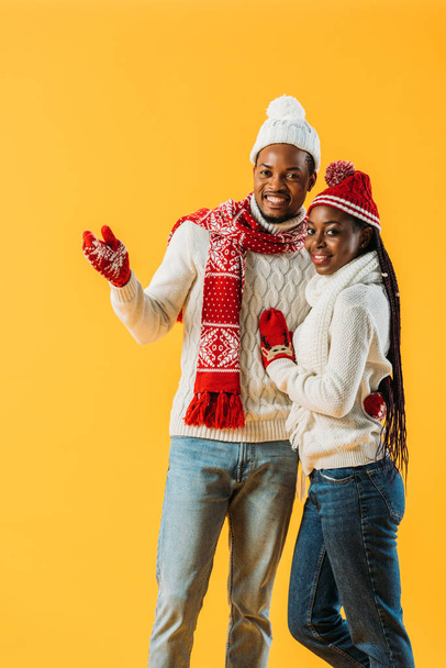 African American man in cozy winter outfit hugging woman and pointing with hand isolated on yellow - Fotoğraf, Görsel