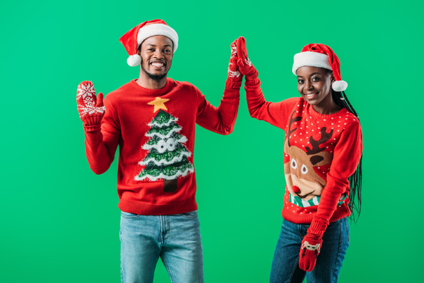 African American couple in red Christmas sweaters and Santa hats giving high five to each other and looking at camera isolated on green - Фото, изображение