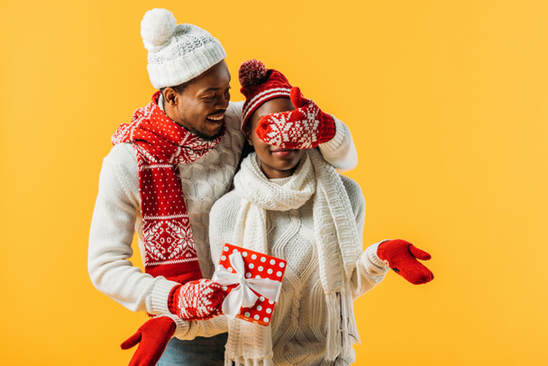 African American man in cozy winter outfit holding gift box and covering with hand eyes of woman isolated on yellow - Foto, Imagem