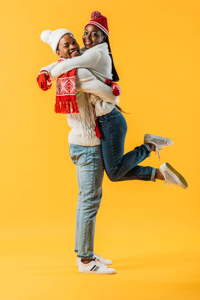 African American man in winter outfit holding up woman isolated on yellow - Foto, immagini