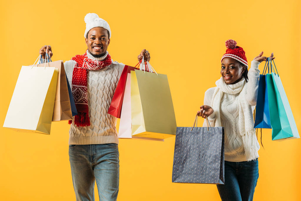 黄色で隔離されたショッピングバッグを持つ冬の衣装でアフリカ系アメリカ人のカップル - 写真・画像