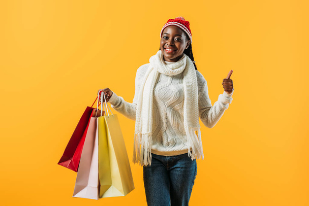 Mujer afroamericana en traje de invierno sosteniendo bolsas de compras y dando pulgar hacia arriba aislado en amarillo
 - Foto, Imagen