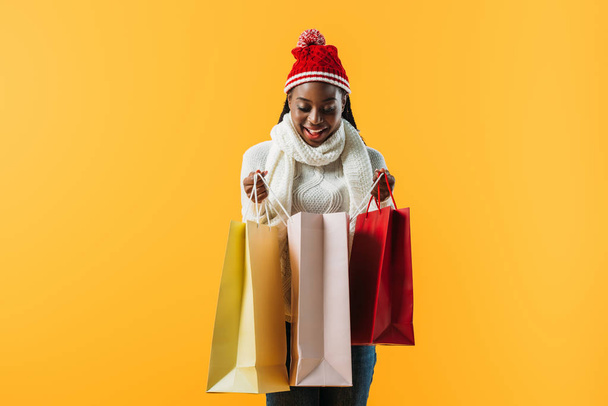 African American woman in winter outfit holding shopping bags and looking inside isolated on yellow - Valokuva, kuva