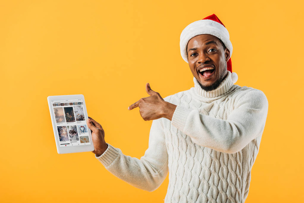 KYIV, UKRAINE - AUGUST 2, 2019: African American man in Santa hat pointing with finger at digital tablet with ebay website isolated on yellow - Fotó, kép