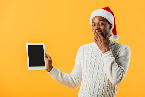 冬のセーターとサンタ帽子のアフリカ系アメリカ人男性は、空白の画面と黄色で隔離された手で口を覆うデジタルタブレットを保持 - 写真・画像