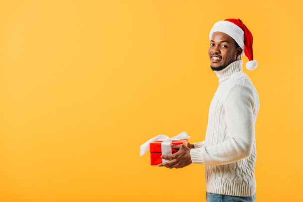 vista lateral del hombre afroamericano en Santa sombrero sosteniendo caja de regalo y mirando la cámara aislada en amarillo
 - Foto, imagen