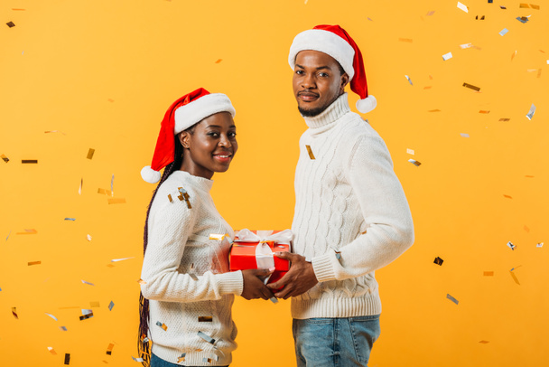 side view of African American couple in Santa hats holding gift box on yellow background with confetti - 写真・画像