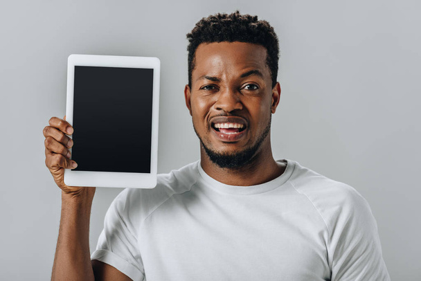 upset African American man holding digital tablet with blank screen and looking at camera isolated on grey - Valokuva, kuva
