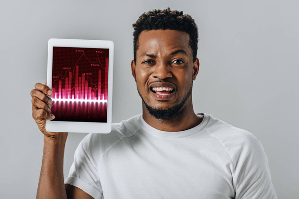 afligido hombre afroamericano sosteniendo tableta digital con gráficos de negocios y mirando a la cámara aislada en gris
 - Foto, imagen