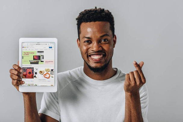 KYIV, UKRAINE - AUGUST 2, 2019: African American man holding digital tablet with ebay website and showing money gesture isolated on grey - Foto, Imagem