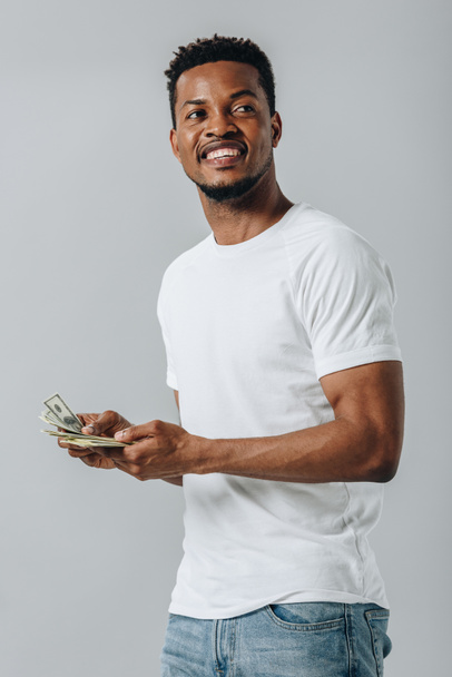 African American mężczyzna trzymający banknoty dolara i patrząc z dala izolowane na szarym - Zdjęcie, obraz