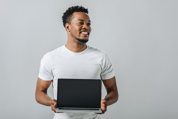 Afro-Américain souriant et montrant ordinateur portable avec écran blanc et regardant loin isolé sur gris
 - Photo, image