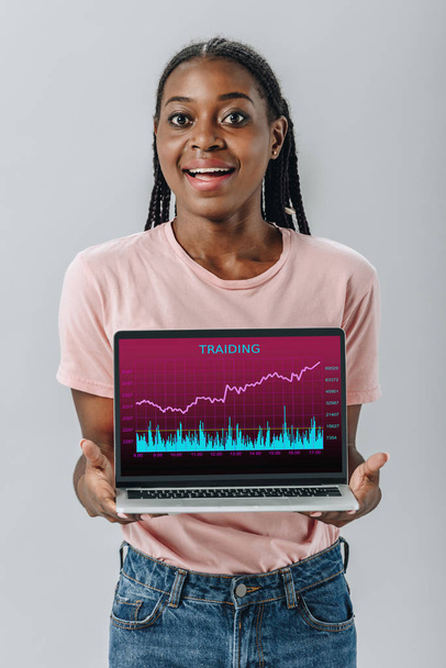 African American woman smiling and holding laptop with trading charts isolated on grey - Fotoğraf, Görsel