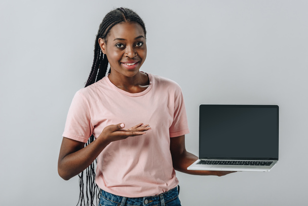 African American woman pointing with hand at laptop with blank screen and looking at camera isolated on grey - Foto, imagen