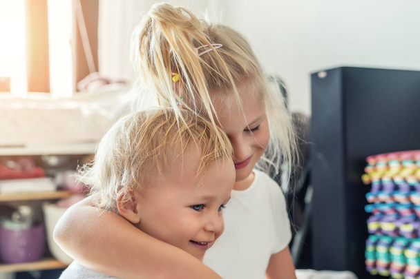 Cute adorable blond caucaian little sister hugging her toddler brother.couple of cheerful sibling playing indoor. Cute girl and baby boy enjoy relaxing at home. Happy childhood, family love, and care - Foto, afbeelding