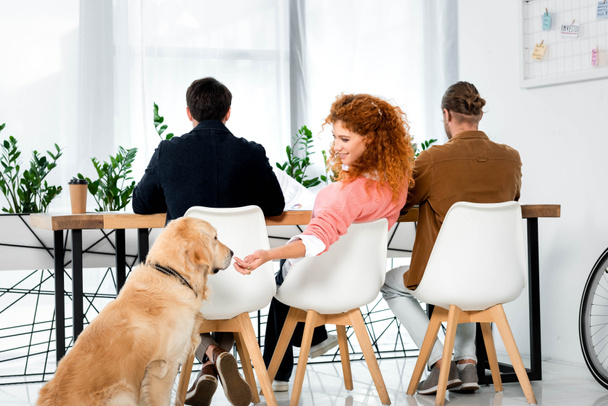 back view of three friends sitting at table and feeding golden retriever - Fotografie, Obrázek