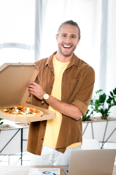 handsome and smiling businessman holding box with pizza in office  - Фото, изображение