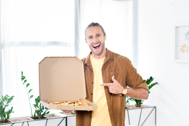 όμορφος και χαμογελαστός επιχειρηματίας δείχνοντας με το δάχτυλο στο κουτί με την πίτσα στο γραφείο  - Φωτογραφία, εικόνα
