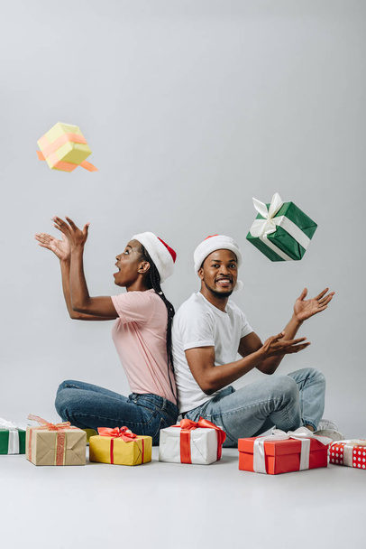 サンタ帽子をかぶったアフリカ系アメリカ人のカップルが後ろに座り、灰色に隔離されたギフトボックスを投げ捨てる - 写真・画像