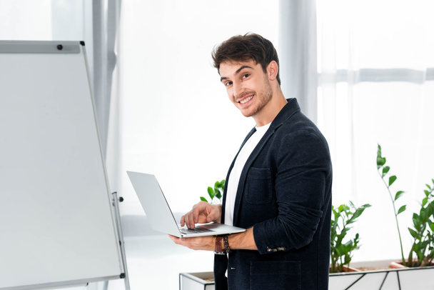 knappe en lachende man in shirt houden laptop en kijken naar camera in Office  - Foto, afbeelding