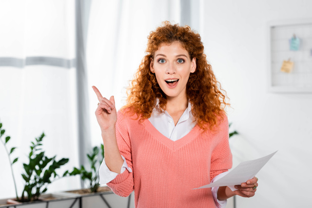 attractive and shocked businesswoman showing idea sign and holding paper  - Photo, Image