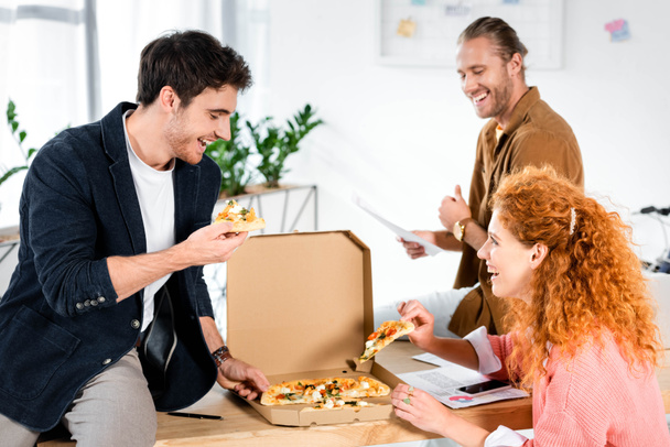 drie lachende vrienden nemen pizza van box in Office  - Foto, afbeelding