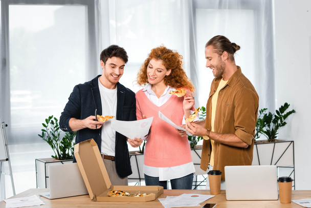 three smiling friends holding pizza slices and doing paperwork in office  - Valokuva, kuva