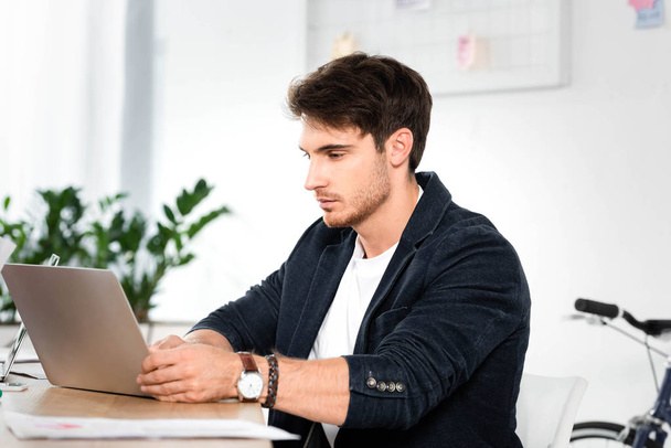 όμορφος επιχειρηματίας σε πουκάμισο χρησιμοποιώντας φορητό υπολογιστή και κάθεται στο τραπέζι στο γραφείο  - Φωτογραφία, εικόνα