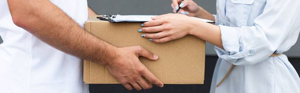 panoramic shot of delivery man holding box near woman signing paper on clipboard outside  - Photo, image