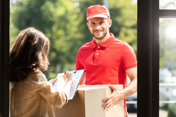 happy delivery man holding box near woman with clipboard and pen  - Photo, Image