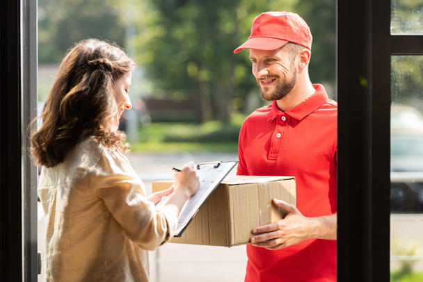 happy delivery man holding box and looking at woman with clipboard and pen  - Φωτογραφία, εικόνα